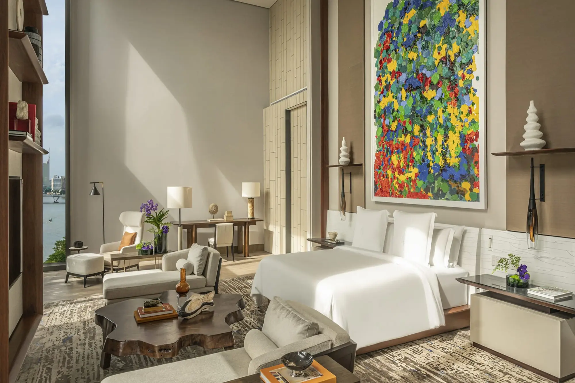 A luxurious bedroom with a large white bed, and large windows offering a water and city view, featuring a colorful abstract painting and elegant furnishings.