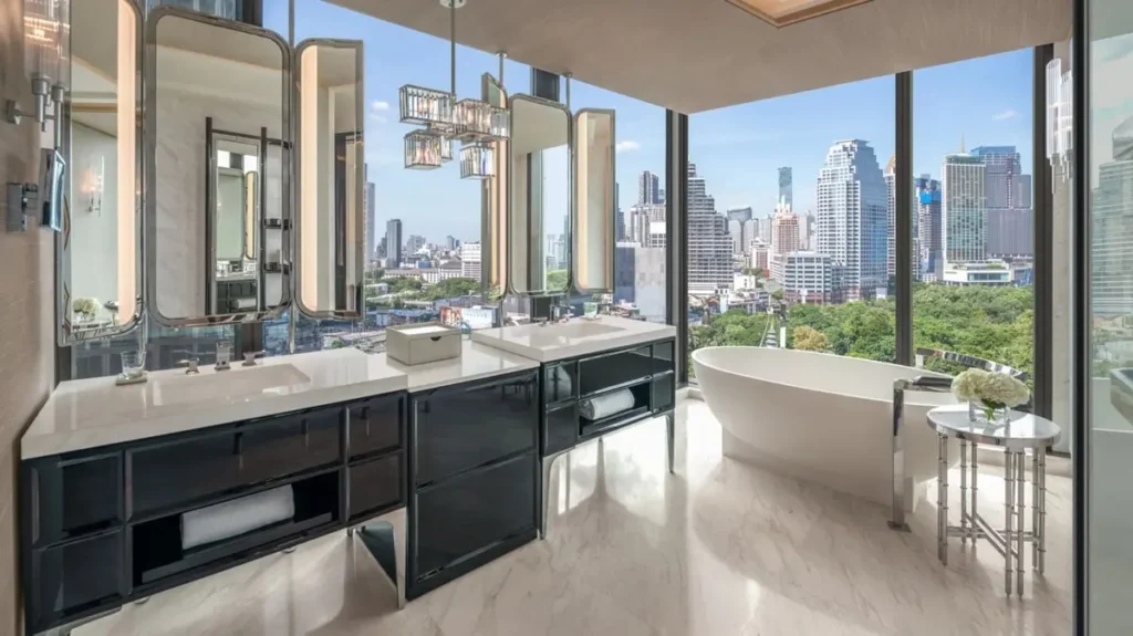 A modern bathroom with large windows showing a city skyline, a double vanity with black cabinets, a white bathtub, chandeliers, and a glass side table with flowers, featuring marble flooring